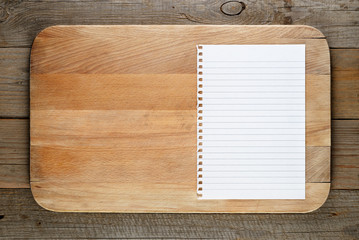 Chopping board and paper for recipe on wooden background