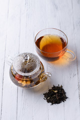 Glass teapot with blooming green tea and cup of tea on white wooden table still life