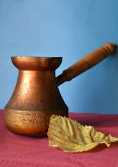 Old copper Turkish coffee pots and yellow autumn leaf on burgundy surface and blue background