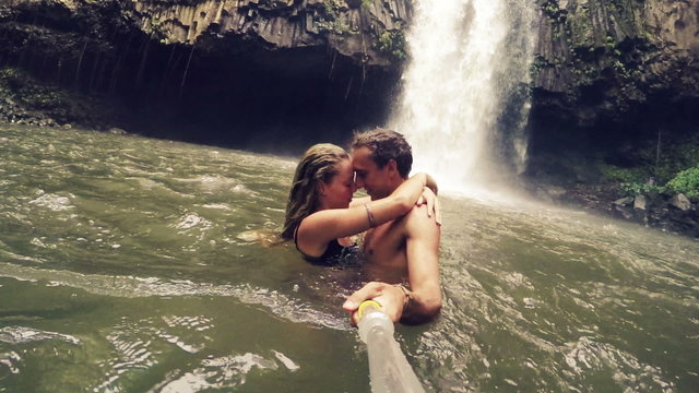 Cute Young Couple Holding Kissing in front of Waterfall with POV Selfie Stick. Happy cheerful young multicultural couple on travel. Lush Green Waterfall in Hawaii.
