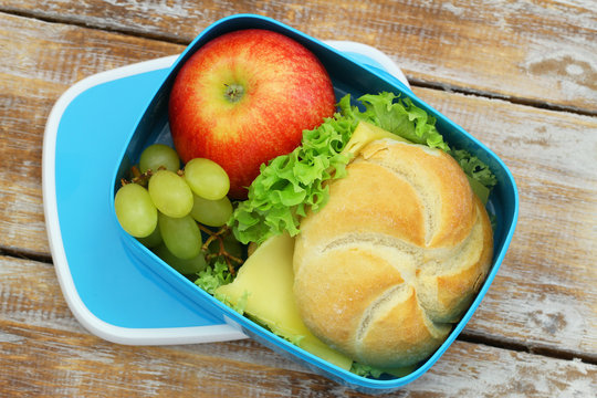 Lunch box containing cheese roll, red apple and grapes on rustic wooden surface

