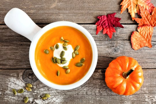 Pumpkin soup topped with pumpkin seeds and cream on rustic wood background with autumn leaves