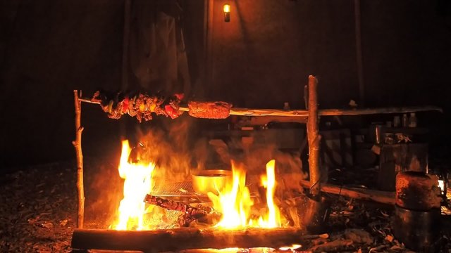 Roasting meat on a wooden pole over a campfire inside a tipi lit by oil lamps. Newly baked Round bread with black crust from a pot is still steaming in the lower right, Cozy primitive cooking