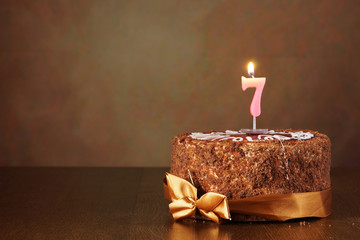 Birthday chocolate cake with burning candle as a number seven on brown background