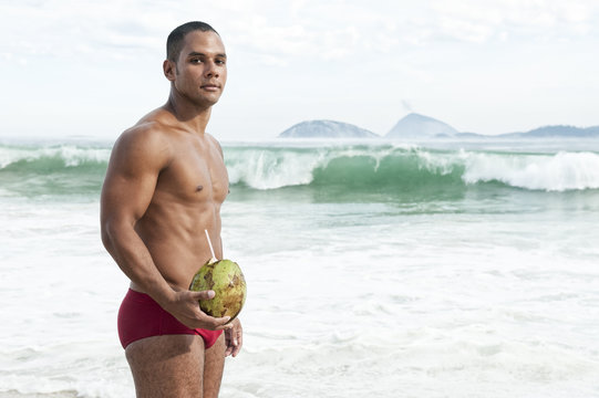 Muscular young carioca Brazilian man wearing red sunga bathing suit holding green coco gelado drinking coconut on Ipanema Beach in Rio de Janeiro, Brazil
