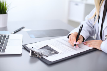 Fototapeta premium Close up of female doctor filling up a medical history form, sitting at the table in hospital