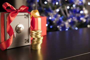 Little safe with red ribbon, golden coins on dark surface. Christmas tree with beautiful decorations and lights on the background