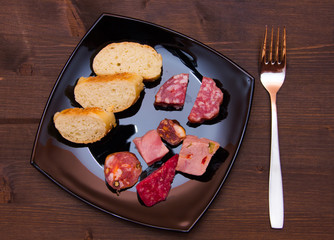 Pieces of salami and bread slices on plate on wooden table seen from above
