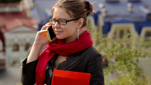beautiful woman in glasses with red folder on the phone against summer buildings
