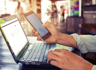 Side view shot of young business man working on his laptop and using smart phone sitting at wooden table in a coffee shop with retro filter effect