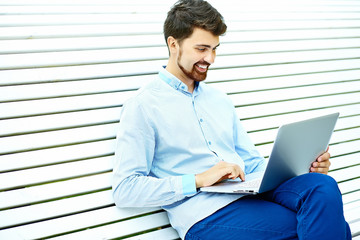 Young handsome smiling businessman model sitting on the park bench using laptop in casual hipster cloth