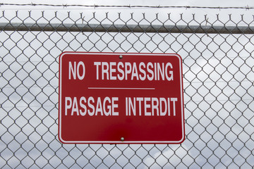 horizontal image of a big red sign saying No Trespassing in english and french language posted on a chain link wire fence with a cloudy sky in background with copy space.
