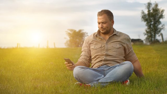 young man looking at smartphone and then throwing it away while sitting outdoors on green grass at autumn and enjoy the nature 