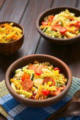 Two rustic bowls of pasta salad made of tricolor fusilli, sweet corn, cucumber, cherry tomato and sausage, photographed with natural light (Selective Focus, Focus one third into the first salad)