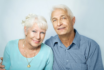  Seniorenpaar sitzen nebeneinander, er hält sie im Arm und beide lächeln in die Kamera 