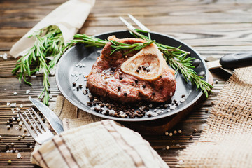 
fried meat in a frying pan with rosemary , salt, pepper and tomatoes on a wooden background