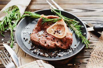 
fried meat in a frying pan with rosemary , salt, pepper and tomatoes on a wooden background