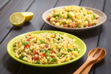 Vegetarian couscous salad with bell pepper, tomato, cucumber, red onion, sweet corn, photographed on dark wood with natural light. (Selective Focus, Focus one third into first salad)