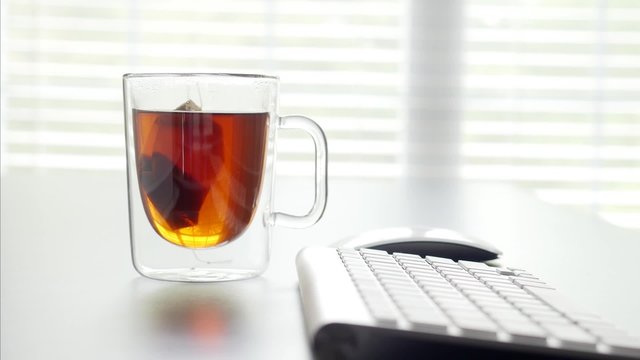 Tea being brewed in a glass cup in an office, transition and zooming in.