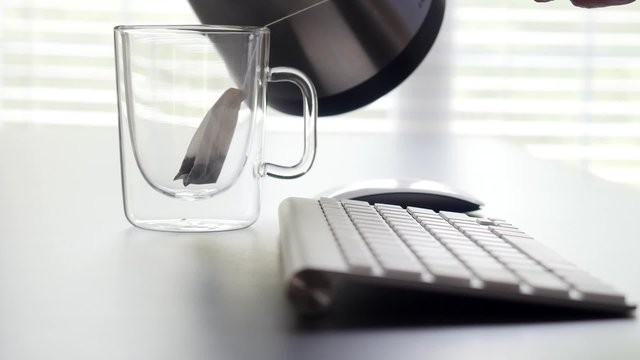 Hot tea pouring into a transparent cup with a teabag in it in an office, zoom in.