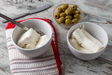 Pezzi di formaggio cremoso in tazza di ceramica bianca e olive verdi snocciolate nella ciotola di su un tavolo di legno