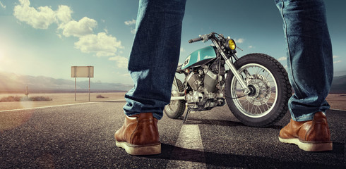 Sport. Biker standing near the motorcycle on an empty road at sunny day. Close view on legs
 - obrazy, fototapety, plakaty