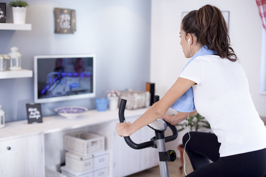 Young sporty woman training on exercise bike in the living room  and watching tv