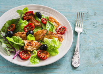Fresh salad with chicken breast, sun-dried tomatoes, green salad and olives on a white plate on a wooden surface. Healthy food