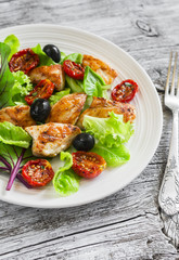 Fresh salad with chicken breast, sun-dried tomatoes, green salad and olives on a white plate on a wooden surface. Healthy food