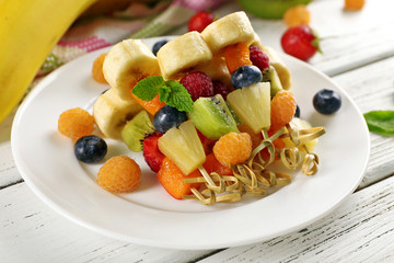 Fresh fruits on skewers in plate on wooden table, closeup