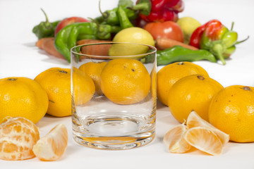 Empty glass cup next to several mandarins and tangerine segments. A bunch of different vegetables at background