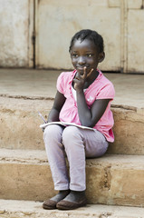 African School Girl concentrated writing notes in her exercise book, lightly smiling sitting on...