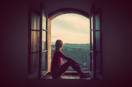 Young woman sitting in an open old window looking on the landscape of Tuscany, Italy.