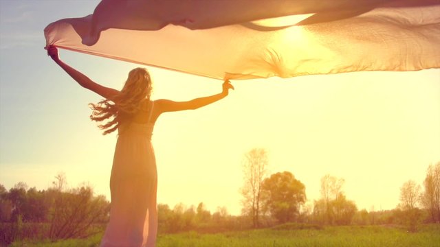 Girl in long dress with chiffon scarf running on summer field and raising hands