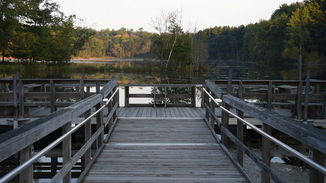 A floating wooden fishing dock sits on the calm waters of pond surrounded by trees, in the early morning.