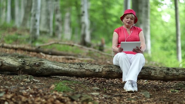 Woman in red hat, T-shirt, white trousers sits on log and talks on skype