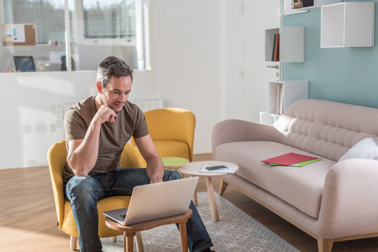 Picture of a middle aged grey hair man with beard in a stylish vintage living room with wooden floor. He is sitting in an orange chair with his laptop in front of him He is wearing casual clothes