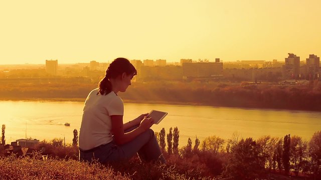 Young woman sitting on the hill with tablet pc in hands. Evening cityscape background 