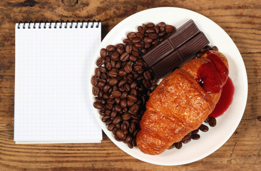 Coffee grains chocolate bars and croissant on white plate beside