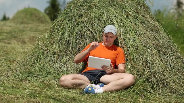 Man in orange t-shirt with tablet PC sits near a haystack