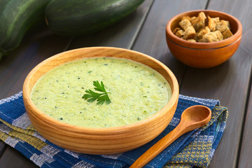 Cream of zucchini soup in wooden bowl garnished with parsley leaf, croutons in the back,...