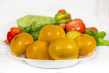 A dish with tangerines in front of a pile of vegetables. On white background