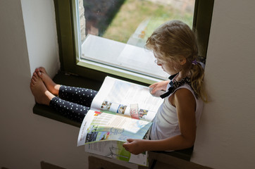 little girl sitting by window and reading brochure
