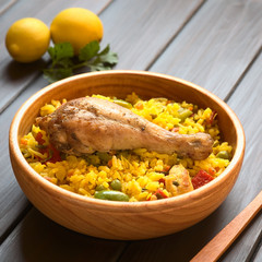 Chicken paella, a traditional Valencian (Spanish) rice dish made of rice, chicken, peas and capsicum , photographed on dark wood with natural light (Selective Focus, Focus on the chicken thigh)