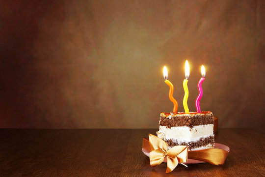 Piece of birthday chocolate cake with burning candles against brown background