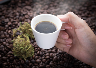 Cannabis coffee. Hand grabbing an ear cup of hot espresso as beside cannabis buds lay on many roasted coffee beans.