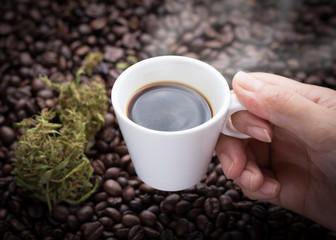 Cannabis coffee. Hand grabbing an ear cup of hot espresso as beside cannabis buds lay on many roasted coffee beans.