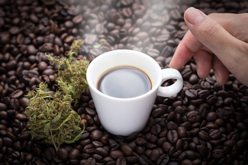 Cannabis coffee. Hand grabbing an ear cup of hot espresso as beside cannabis buds lay on many roasted coffee beans.