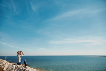 Girl with binoculars at the mountains in the rays of the evening sunset. Young beautiful woman on nature background. She is resting in the open air. Keep holding binoculars. Look ahead. Copy space