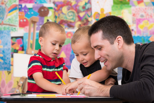 Happy young father with his two little sons painting something, smiling and having a good time. Colorful wall with paintings and easel in background.